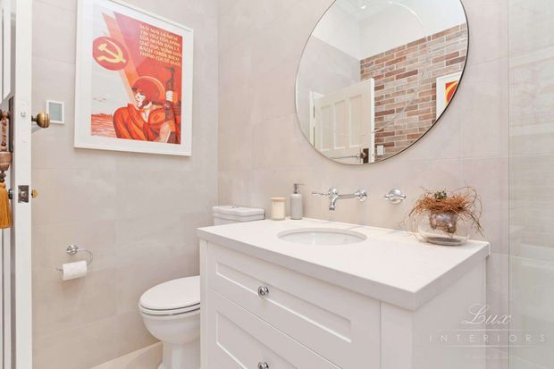 A compact, bright bathroom with pale tiled walls, a white vanity with paneled drawers and an integrated basin, wall-mounted chrome tapware, a toilet, and a large round mirror. The mirror reflects a partially open white door and a brick-patterned wall. A framed red vintage-style poster adds a bold decorative accent.