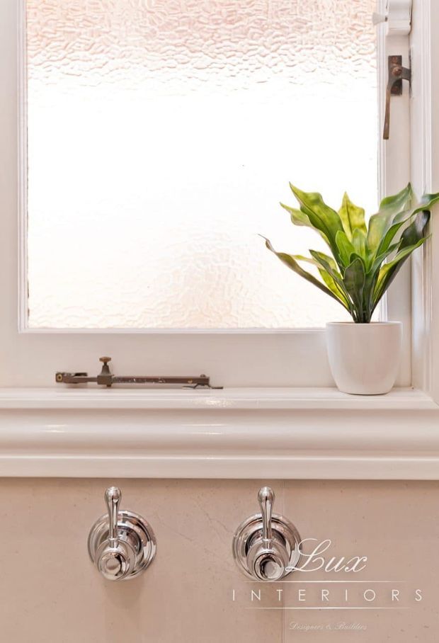 Close-up interior detail of a traditional-style bathroom or washroom window with frosted textured glazing, white painted trim and sill, chrome plumbing fixtures below, and a potted green plant. The image highlights refined period-inspired detailing and polished hardware.