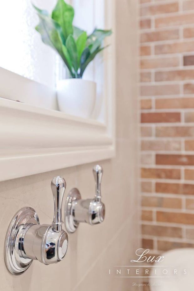 Close-up detail of a refined bathroom wall featuring polished chrome plumbing fixtures, a pale stone or tile wall finish, white trim and ledge, a partially visible bathtub, and a potted plant on the windowsill. A warm red brick feature wall is visible to the right, creating contrast with the light neutral finishes.