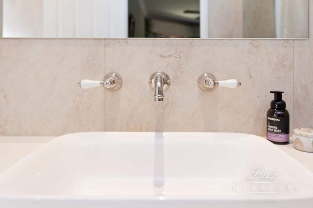 Close-up view of a refined bathroom vanity with a white ceramic basin, wall-mounted chrome tapware, pale beige stone or porcelain wall tiling, and a large mirror above. The styling is clean and understated with light neutral finishes.