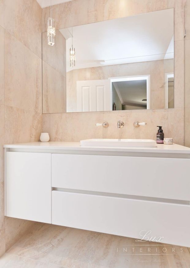 Bright, refined bathroom featuring a wall-mounted white vanity with integrated basin, minimalist chrome wall-mounted tapware, large frameless mirror, and pale beige stone-look tiled walls and floor. Decorative pendant lights and a reflected doorway add detail, while the overall scheme is clean and understated.