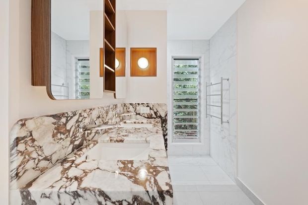 Bright contemporary bathroom featuring a floating vanity with a dramatic veined stone benchtop and matching splashback, warm timber-framed mirrored cabinetry, white marble-look wall and floor tiles, louvred windows, and a wall-mounted towel rail.