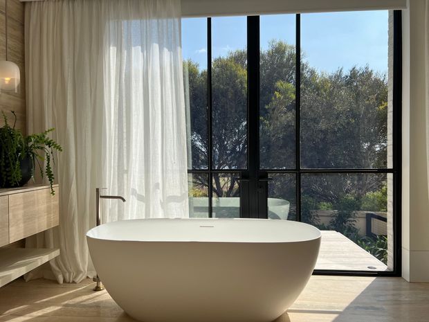 A serene, light-filled contemporary bathroom featuring a freestanding oval bathtub positioned in front of large black-framed floor-to-ceiling glazing. Sheer curtains, warm timber wall and floor finishes, and a floating timber vanity with integrated shelving create a calm, refined atmosphere. A leafy outdoor setting is visible beyond the windows.