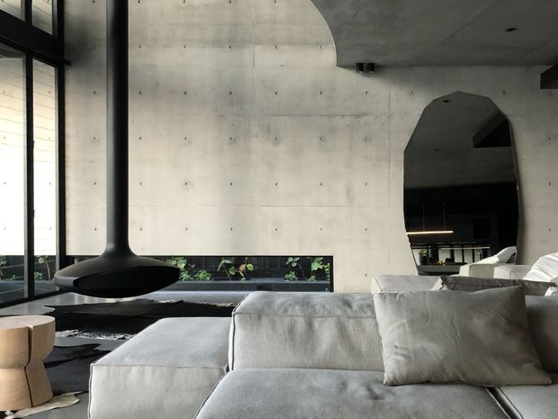 A spacious contemporary living room with a large pale sectional sofa in the foreground, a sculptural suspended black fireplace, exposed smooth concrete walls and ceiling, full-height black-framed glazing, and a large irregularly shaped wall mirror reflecting the seating area. A narrow planted lightwell runs beneath the windows, adding greenery to the otherwise monochromatic interior.