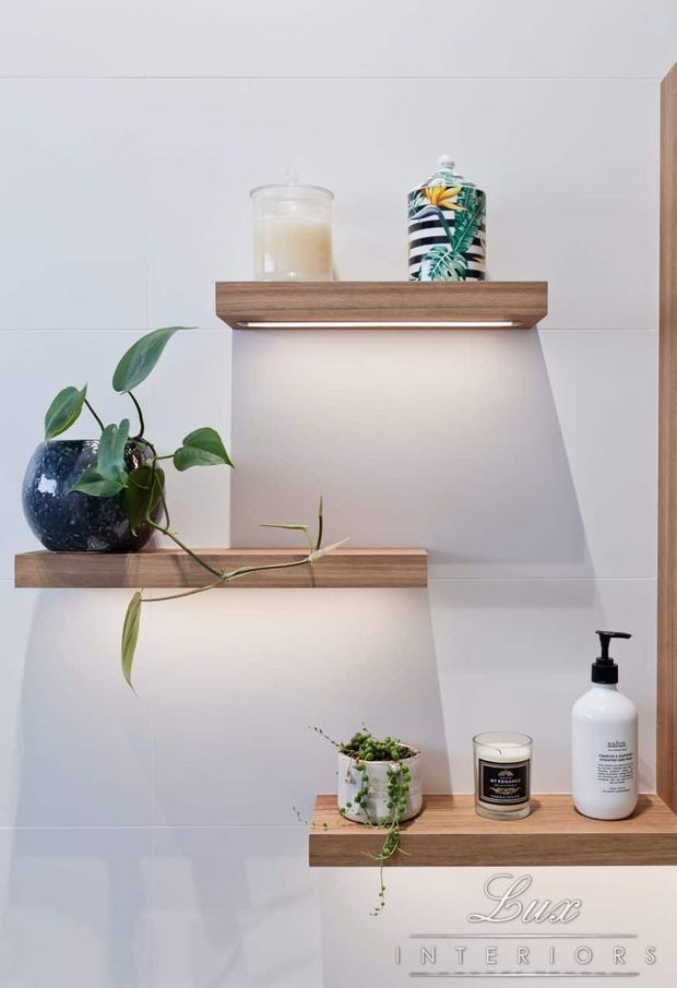 A close-up view of a white tiled wall with three staggered floating timber shelves. The shelves display potted trailing plants, candles, a decorative patterned jar, and a bathroom product dispenser. Integrated strip lighting beneath the upper shelves creates soft illumination, while a timber vertical edge frames the right side.