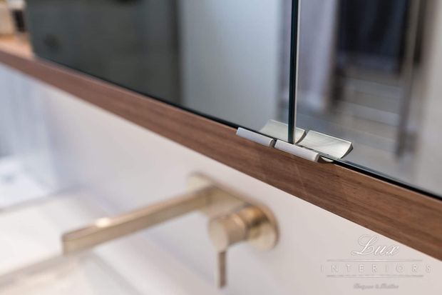 Close-up detail of a contemporary bathroom vanity or basin area, showing a frameless mirror with a dark timber-toned lower frame or shelf, polished metal mirror hardware, and a wall-mounted brushed metal tap against a white wall. The image has a refined, minimalist luxury interior presentation.