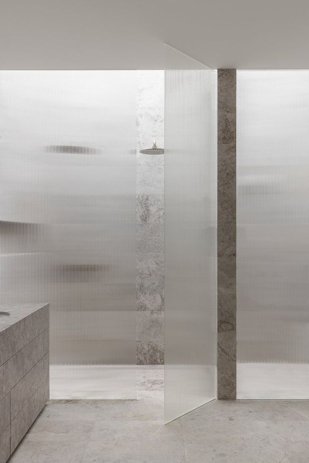 A minimalist contemporary bathroom shower area with translucent vertically ribbed glass partitions, pale stone wall and floor tiles, a frameless glass enclosure, and a wall-mounted showerhead. The space uses a restrained light grey and white palette with subtle stone veining and clean geometric lines.