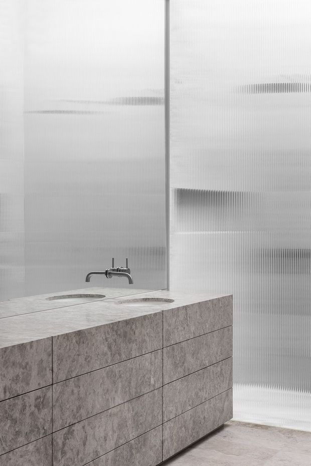 Minimalist contemporary bathroom featuring a double vanity clad in grey stone-look panels with integrated basins and a wall-mounted brushed metal tap. Translucent vertically ribbed glazing forms the backdrop, creating a soft, diffused and highly textured enclosure. The floor and vanity use coordinated grey stone finishes.