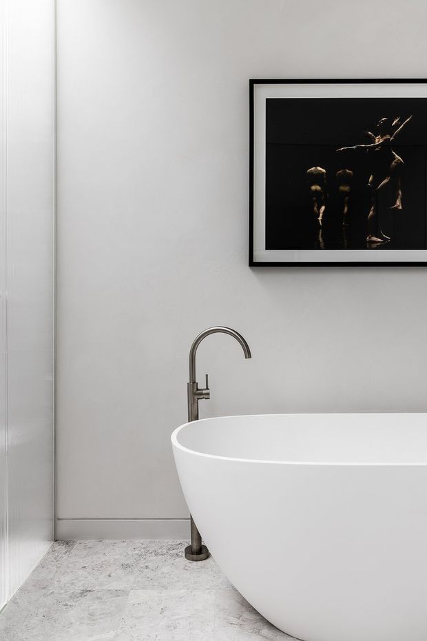 A minimalist bathroom featuring a white freestanding bathtub, brushed dark metal floor-mounted gooseneck tap, pale grey wall, and light marble-look tiled flooring. A framed dark artwork hangs above the bath, while a frameless glass shower enclosure is visible at the left edge.