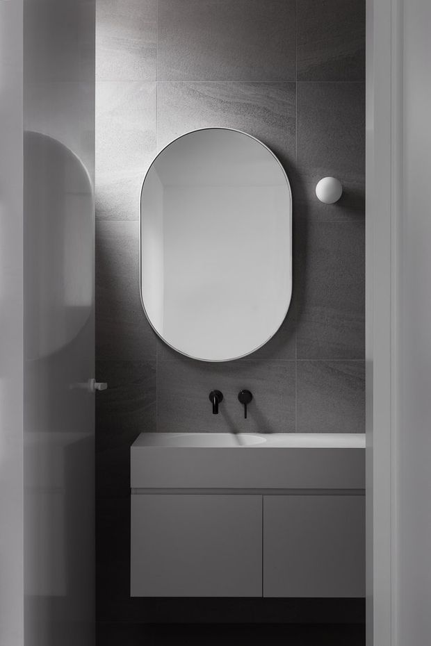 A compact contemporary bathroom viewed through a doorway, featuring a floating white vanity with integrated basin, dark grey tiled walls, black wall-mounted tapware, a rounded-rectangle mirror, and a spherical wall light. The restrained monochrome palette and clean lines create a minimalist, refined atmosphere.