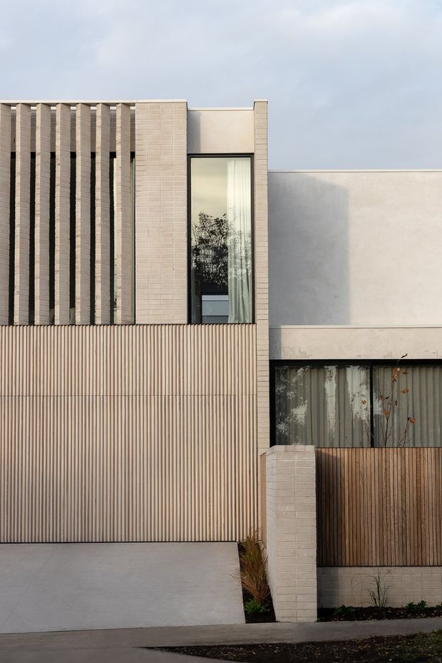 A close exterior view of a contemporary multi-storey residence with a restrained neutral palette. The facade combines pale masonry or tiled vertical fins, a tall narrow window, smooth rendered wall planes, vertically ribbed cladding, timber-toned screening, and recessed glazing. A concrete driveway and planted entry strip sit in the foreground.