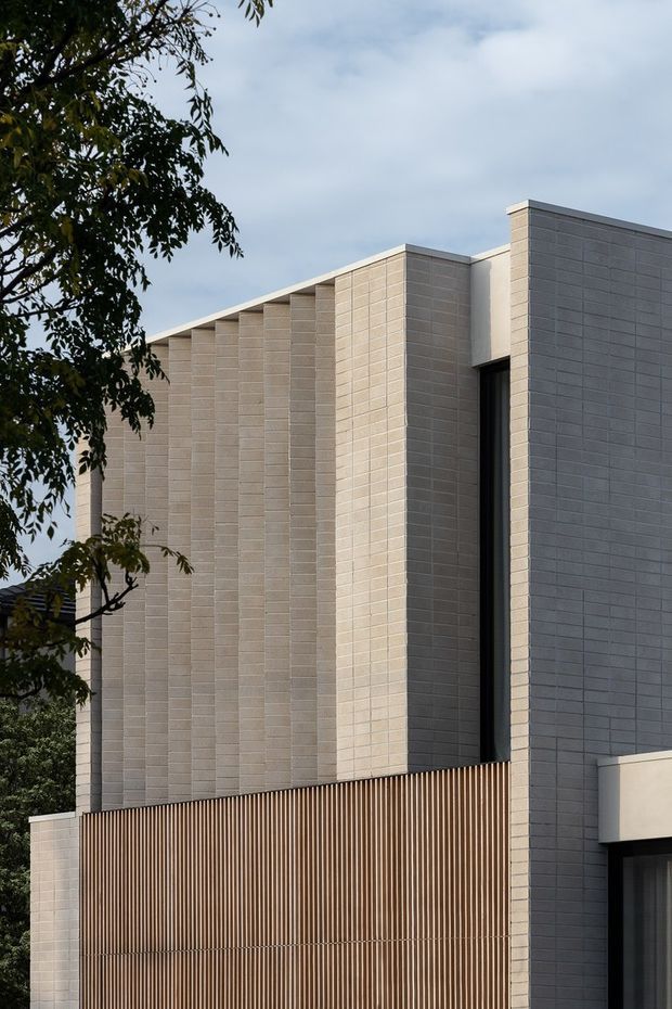 Close-up exterior view of a contemporary multi-storey building with a strong geometric facade. Pale grey and warm beige masonry or rendered blockwork forms repeated vertical fins and projecting wall planes, contrasted by a tall narrow dark window opening and a brown vertical timber-look privacy screen. Trees partially frame the building at left.