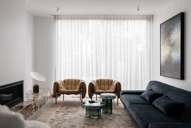 A refined contemporary living room with a large sheer-curtained window, dark sectional sofa, two tan leather lounge chairs, glass coffee tables, a marble-clad fireplace, and neutral abstract artwork. The space is bright, calm, and softly layered with warm neutral textures.