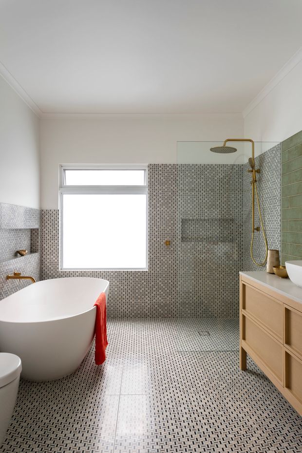 Bright contemporary bathroom featuring a freestanding white bathtub, walk-in shower with frameless glass, patterned mosaic wall and floor tiles, a green tiled shower feature wall, brass shower fittings, and a light timber vanity with a white basin. A window provides natural light, while a red towel adds contrast.