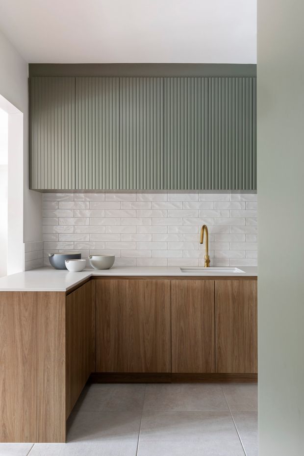 A compact contemporary kitchen with warm timber cabinetry, a pale stone or composite benchtop, white textured subway-tile splashback, and a vertically fluted grey-green overhead cabinet. A slim brass/gold tap and undermount sink sit beneath the upper joinery. Large pale floor tiles and soft neutral walls create a clean, refined, minimalist composition.