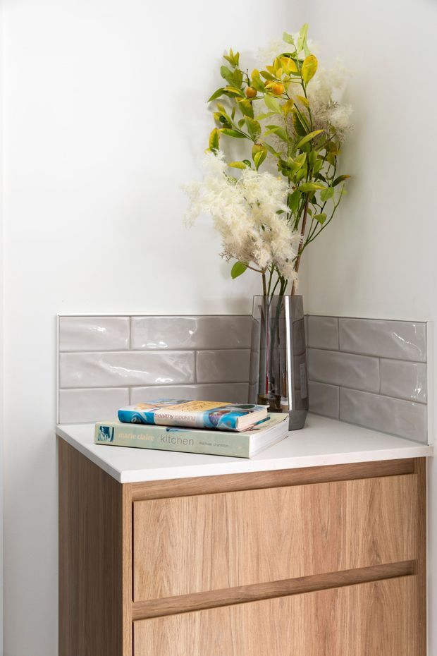 Close-up interior detail of a light timber built-in cabinet or vanity with a white benchtop and glossy pale grey subway-tile splashback. A tall reflective metal vase with leafy branches and white dried flowers sits on the surface beside stacked books. The composition is bright, neutral, and softly styled.