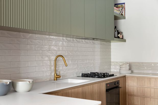Contemporary kitchen featuring pale timber cabinetry, a white engineered benchtop, glossy white subway-tile splashback, vertically fluted sage-green upper cabinetry, and a brass-gold gooseneck tap. Integrated gas cooktop and oven sit beside open shelving with crockery and bottles.