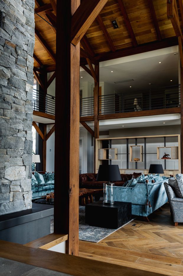 A dramatic double-height living room with exposed dark timber beams and a pitched timber ceiling, a substantial stacked-stone feature wall and fireplace, black metal balustrades, and patterned timber parquet flooring. The room is furnished with blue velvet sofas, leather seating, dark side tables, lamps, and built-in shelving, with a mezzanine level overlooking the space.