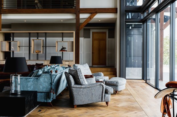 A spacious double-height living room with a mezzanine level, exposed dark timber structural beams, full-height black-framed glazing, and a warm herringbone timber floor. The room contains a large blue sectional sofa, grey upholstered armchairs and ottomans, a black coffee table, and built-in open shelving. A timber-clad entry door and staircase are visible at the rear, creating a refined contemporary interior with strong natural materials and abundant daylight.