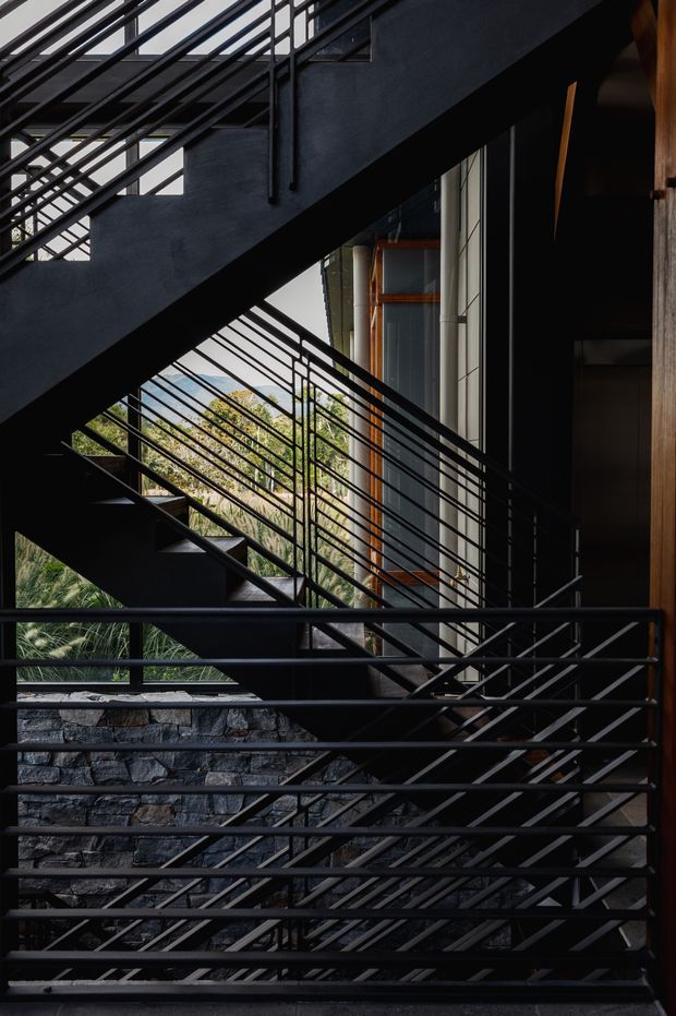 A dramatic architectural view of a multi-level stairway with black steel stringers and tightly spaced metal railings. The stairs are integrated into a dark, textured building facade beside tall glazing and warm timber-framed openings. Layered stair flights, exposed structural geometry, stone retaining walls, and dense greenery visible beyond create a strong indoor-outdoor composition with high contrast.