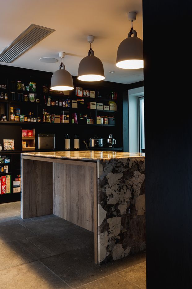 A dark, refined kitchen or pantry space with extensive black open shelving stocked with food and kitchen supplies. A compact island features timber-look panelled cabinetry, a dramatically patterned natural-stone waterfall end panel, and a light stone benchtop. Three oversized pendant lights hang above the island, while dark tiled flooring and black walls create a moody, high-contrast interior.