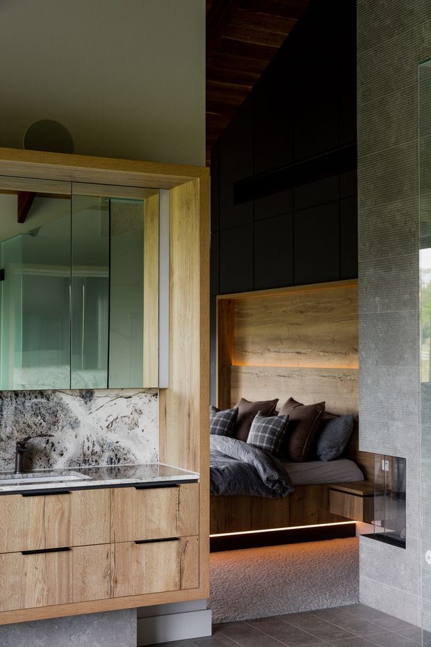 A sophisticated compact bedroom suite viewed through a bathroom or vanity area. The room features warm timber cabinetry and wall panelling, a dark built-in bedhead with integrated amber lighting, layered charcoal and checked bedding, a grey carpeted sleeping platform, dark tiled flooring, and a freestanding or built-in vanity with mirrored upper cabinetry and a dramatic pale stone splashback. A textured grey screen or wall element partially divides the spaces. The design is moody, refined, and highly integrated with concealed lighting and custom joinery.