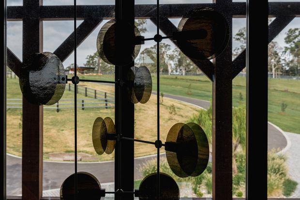 A large glazed architectural opening viewed from inside, featuring a suspended sculptural installation of dark circular discs and metal rods in front of the glass. The glazing is framed by dark timber or metal structural members, with a landscaped rural setting and winding paved path visible beyond.