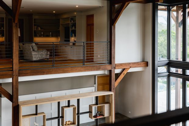 Interior view of a multi-level living space with a mezzanine overlooking a lower level. Exposed dark timber posts, beams and diagonal braces frame the space, with metal cable railings, built-in shelving, warm timber flooring and large floor-to-ceiling windows. A partially visible lounge and open-plan kitchen or living area sit on the upper level, while decorative built-in shelving occupies the lower level.