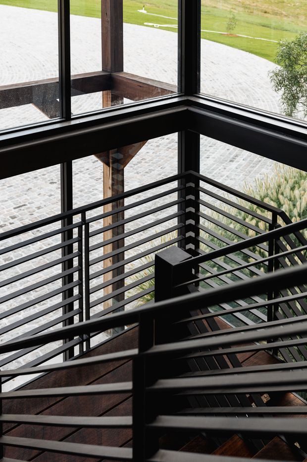 Interior view of a stairway with dark timber steps, black metal horizontal balustrades, exposed timber structural posts and large black-framed corner glazing. A paved exterior courtyard and landscaped grounds are visible beyond the windows.