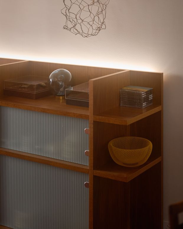 Close-up interior detail of a warm timber storage cabinet or sideboard with open shelving, ribbed translucent sliding or inset panels, curated books and decorative objects, and integrated warm backlighting. A sculptural wire pendant hangs above, creating a refined mid-century-inspired atmosphere.