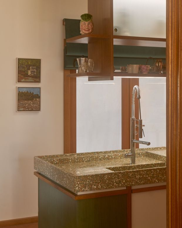 A compact, richly detailed kitchen or kitchenette viewed through a doorway, featuring a dark timber island with a thick terrazzo-style benchtop and integrated sink. The sink has a tall coiled stainless-steel gooseneck tap. Behind it are warm timber-framed open shelves and cabinetry, ribbed translucent glass doors, and a pale reflective splashback. Framed artwork and displayed glassware add a curated, vintage-inspired character.