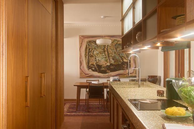 Warm, richly timber-lined kitchen viewed toward an adjoining dining area. The kitchen features extensive built-in wood cabinetry, open shelving, a long stone benchtop with integrated cooktop and sink, pendant and under-cabinet lighting, and a large timber-framed glazed opening. A dining table, patterned rug, and artwork are visible beyond.