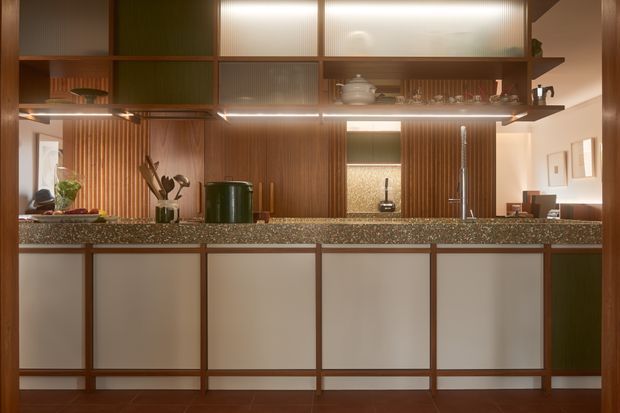 A warm, carefully detailed kitchen viewed straight on from the living side. The room features a long island or peninsula with dark speckled stone benchtops and pale inset cabinet fronts framed in timber. Behind it, vertically grooved timber wall panelling and open shelving support a bank of upper cabinetry with translucent ribbed-glass doors, integrated lighting, and dark green accents. A sink and tap sit along the rear bench, with cookware and small appliances visible. The overall composition has a strong mid-century-inspired, crafted joinery character with warm ambient lighting.