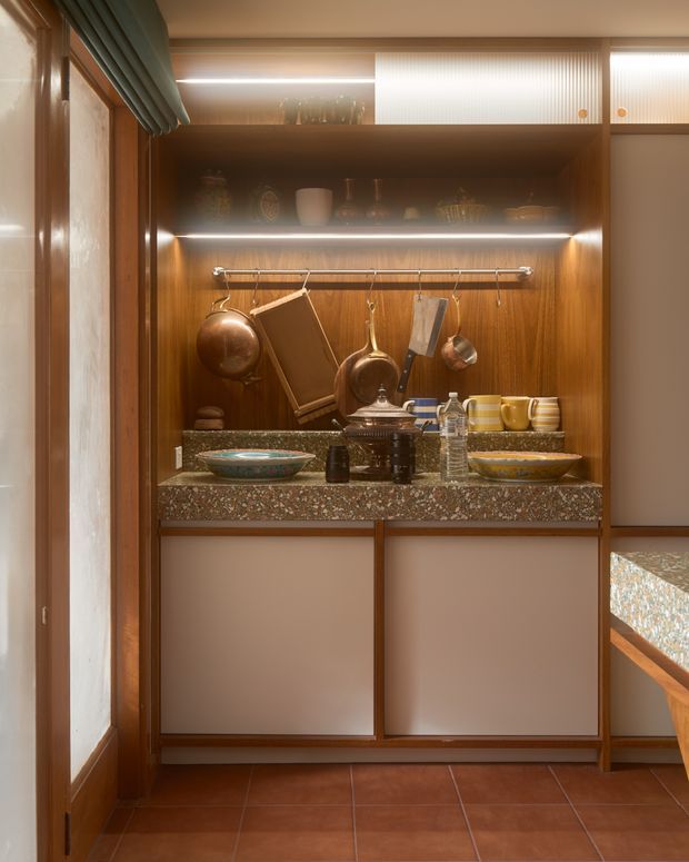 A compact, warm-toned kitchen or butler's pantry with dark timber open shelving and cabinetry, integrated warm LED strip lighting, a speckled stone benchtop, and hanging copper cookware. Crockery and kitchenware are displayed on the open shelves, with a tiled floor and a partial view of an adjoining benchtop at right. The design has a refined mid-century-inspired character with strong timber detailing and retro materials.
