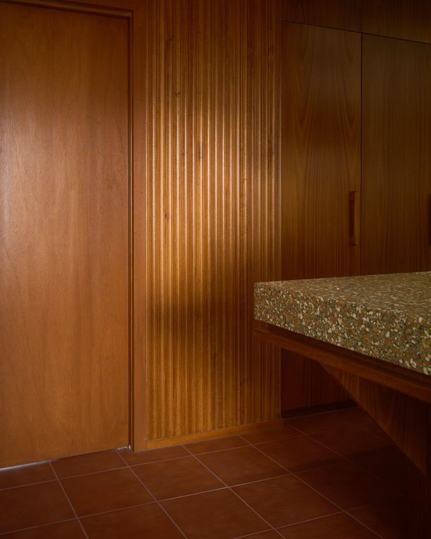 A warmly lit, wood-panelled interior featuring a full-height timber door at left, a vertically fluted timber feature wall in the centre, and dark timber cabinetry at right. A thick terrazzo countertop projects into the foreground, supported by built-in timber joinery. The floor is finished in reddish-brown square tiles, creating a rich mid-century-inspired material palette.