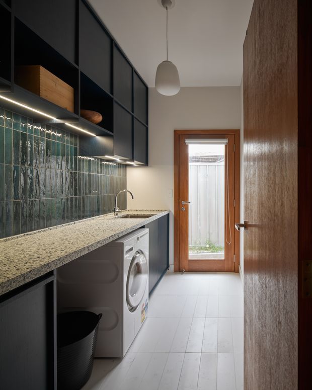 A narrow, well-appointed laundry room with dark built-in cabinetry, a long pale stone benchtop, deep green textured tile splashback, integrated sink, front-loading washing machine, and timber-framed glazed door. Warm under-cabinet lighting and a simple pendant create a refined, functional interior.