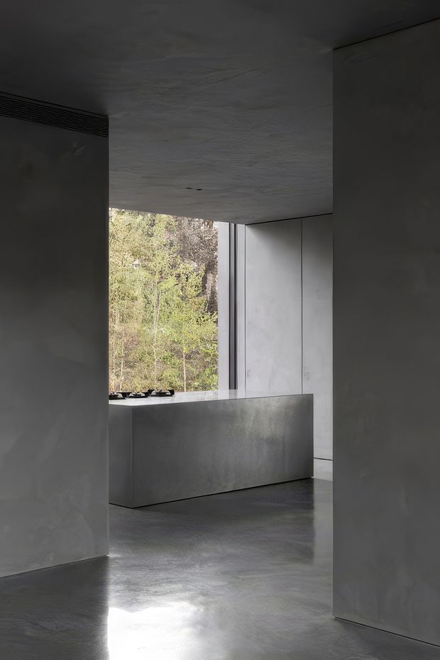 A dark, minimalist interior focused on a sculptural monolithic kitchen island or counter. Smooth grey concrete-like walls, ceiling and floor create a seamless restrained envelope, with flush full-height grey cabinetry and a large vertical window looking onto trees. The space is sparsely furnished, with strong natural light reflected across the polished floor and a highly pared-back contemporary atmosphere.