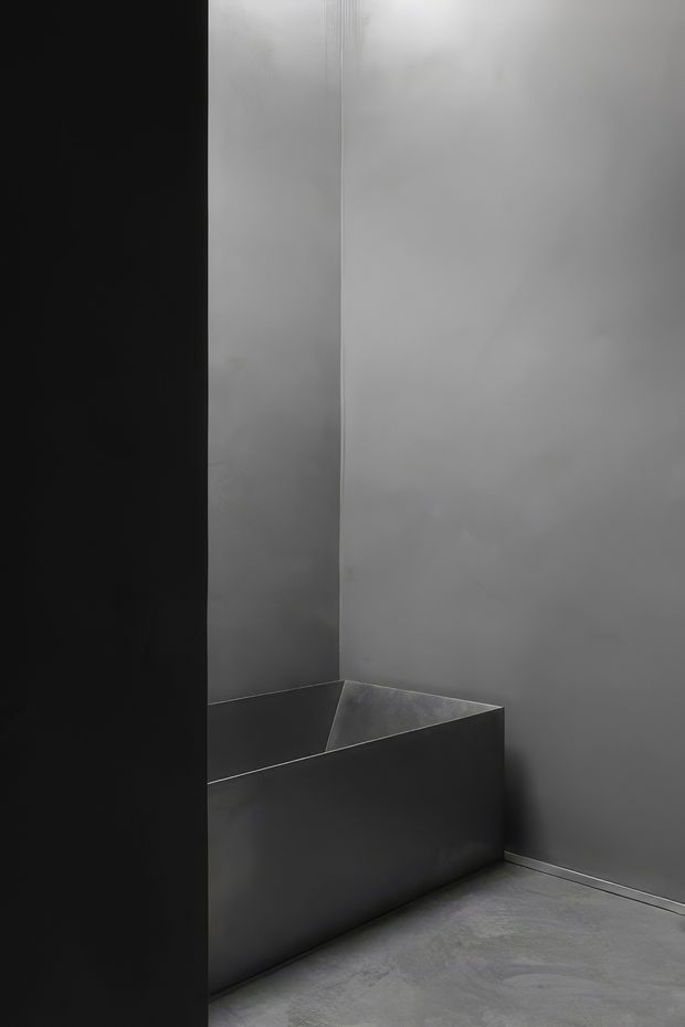 A stark monochromatic interior corner featuring a deep, angular built-in or freestanding dark bathtub against smooth grey walls. The space has a minimal, sculptural character with a seamless grey concrete-like floor and very limited detailing.