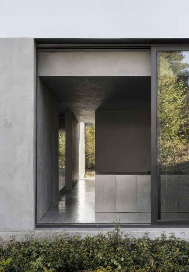 A close exterior view of a minimalist modern building facade, focused on a large dark-framed glazed opening. Through the glazing is a deeply recessed, shadowed passage or interior space with smooth grey walls, a dark ceiling, reflective flooring, and a built-in grey tiled or paneled volume. Trees are visible beyond the passage, while low planting runs along the base of the facade. The composition emphasizes strong geometric lines, deep reveals, and a restrained monochromatic material palette.