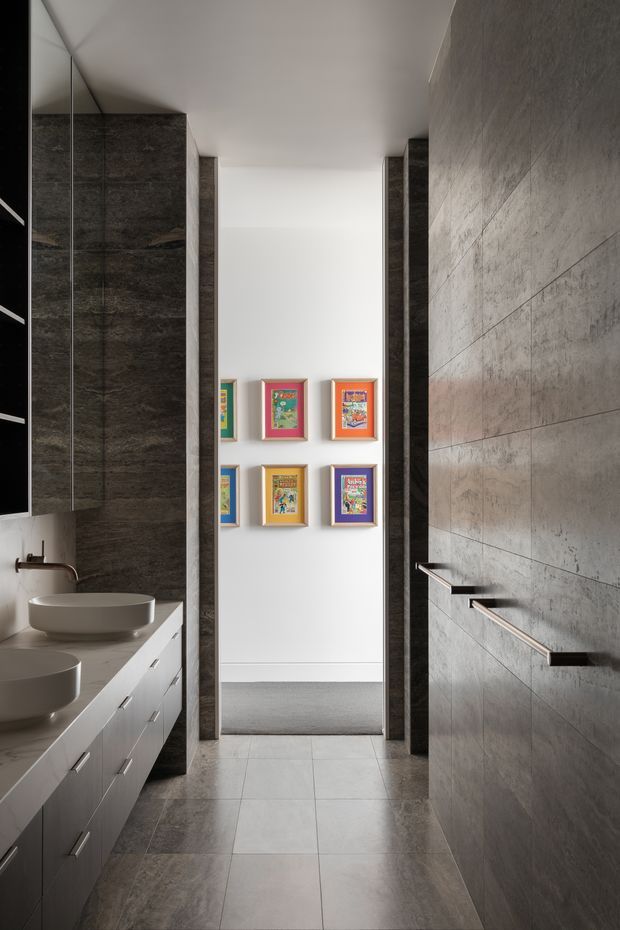 A narrow, sophisticated bathroom with a double vanity at left, dark stone-look cabinetry and wall surfaces, large grey floor tiles, and a long tiled wall with sleek horizontal handles at right. A bright hallway or adjoining room with colourful framed artwork is visible beyond the bathroom.