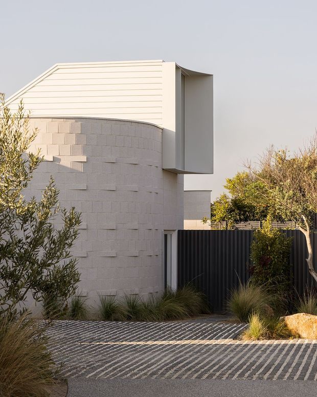 Contemporary residential exterior viewed from the street, featuring a curved pale masonry wall with projecting blocks, horizontal cladding, a sculptural upper volume, dark vertical metal fencing, and a patterned paved driveway. Drought-tolerant grasses and shrubs soften the minimalist facade.