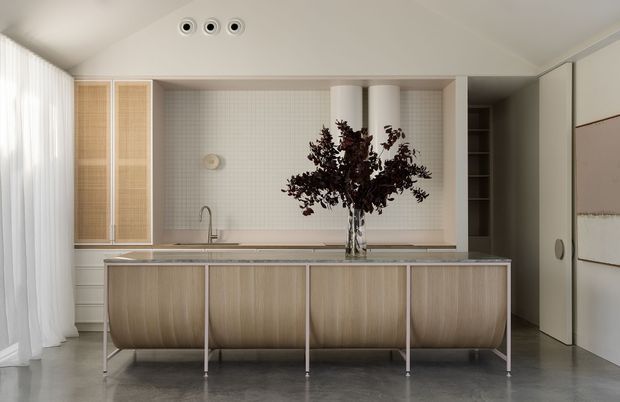 A refined contemporary kitchen with a long island featuring curved light-wood cabinetry and slim metal legs. The space has a pale tiled splashback, stone-look benchtop, integrated sink and tap, tall timber-framed cabinetry with woven or ribbed fronts, concealed storage, sheer curtains, and polished grey flooring. A dark leafy arrangement in a glass vase provides contrast against the soft neutral palette.