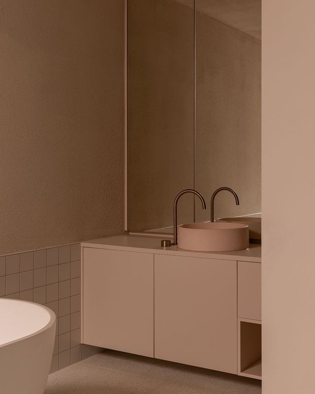 Warm monochromatic contemporary bathroom with a freestanding bathtub partially visible in the foreground, a floating pale pink vanity with integrated storage, a round vessel basin, and twin dark curved faucets. Large mirrored wall panels amplify the compact space, while small square wall tiles and a textured plaster wall add subtle material contrast.