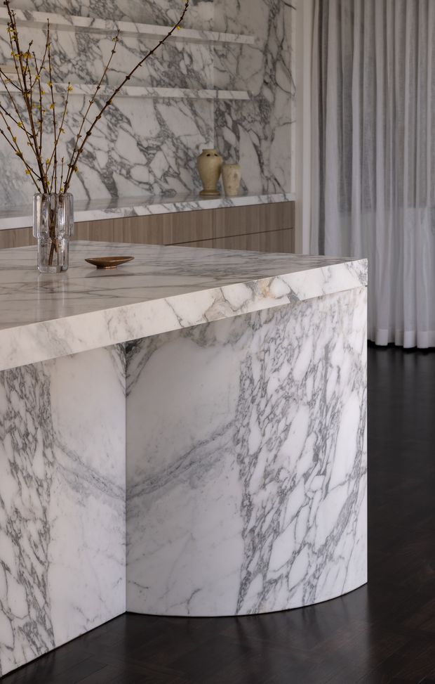 Refined interior detail featuring a sculptural marble kitchen island or counter with a curved end and waterfall sides. Matching bookmatched white-and-grey veined marble forms the backsplash and counter surface, with understated wood cabinetry behind. A glass vase with branches and ceramic vessels add minimal decorative accents, while sheer curtains and dark timber flooring complete the elegant setting.