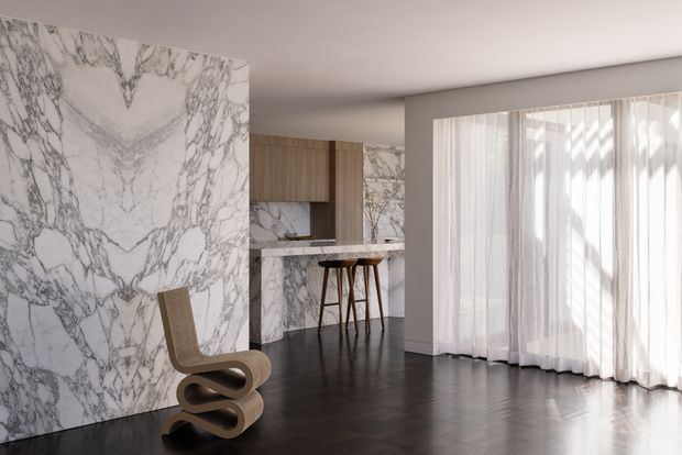 A refined contemporary open-plan interior with dramatic bookmatched white-and-grey veined stone panels forming a feature wall and kitchen or bar surround. Warm timber cabinetry and a slim stone counter sit behind a sculptural wooden bar stool. Dark timber flooring contrasts with the pale walls and full-height sheer curtains, creating a bright, polished and luxurious atmosphere.