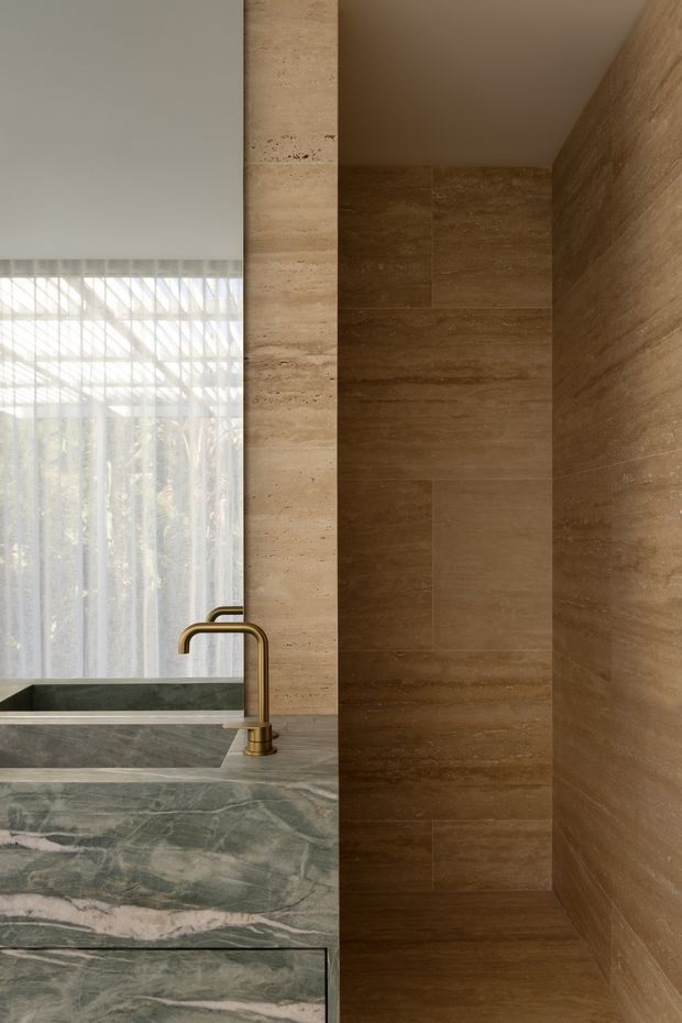 A refined bathroom interior featuring a freestanding or built-in dark stone bathtub beside a sculptural green-grey marble vanity with an integrated basin and brushed brass tapware. Warm brown travertine-like stone tiles line the shower enclosure and adjacent wall, while sheer curtains provide soft filtered daylight. The palette is warm, earthy and luxurious, with strong natural stone detailing.