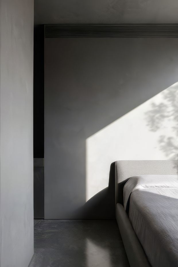 A minimalist bedroom corner with a low upholstered bed in pale grey, set against smooth grey walls and a dark polished concrete floor. Strong sunlight enters from the right, casting a sharp geometric beam and leafy shadow across the wall and floor. A dark linear ventilation grille and black vertical trim provide subtle architectural contrast.