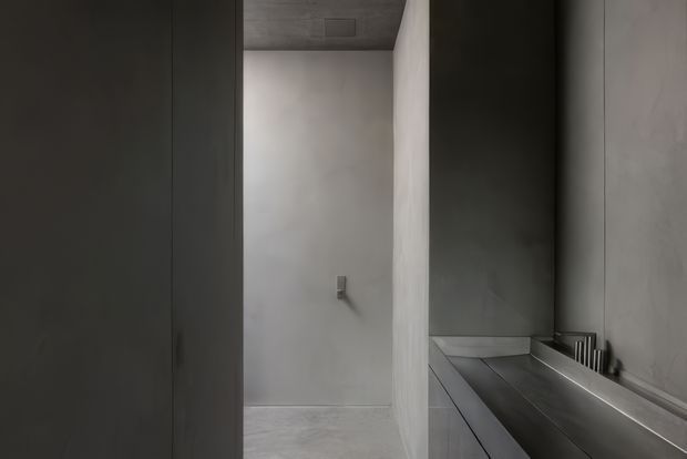 A minimalist monochromatic bathroom interior, viewed toward a walk-in shower with a wall-mounted fixture. A long dark vanity with an integrated basin and taps occupies the right side, while tall dark cabinetry or wall panelling frames the left. Smooth plaster or concrete-like grey surfaces, a matching ceiling, and pale grey flooring create a restrained, seamless appearance.
