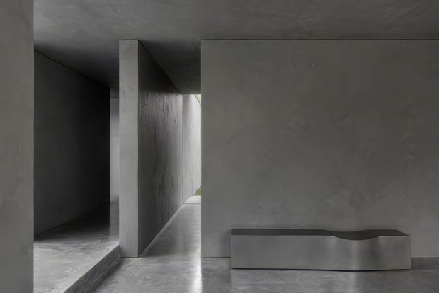 A moody minimalist interior composed of raw grey concrete or plaster surfaces. The view looks through a narrow architectural passage with strong geometric walls, a low ceiling, and a small opening bringing in natural light. In the foreground is a simple sculptural grey built-in or console-like element with a subtly curved top edge. The space is largely empty, monochromatic, and defined by its austere materiality and dramatic shadow.
