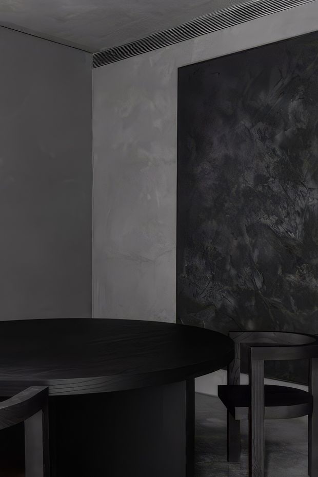 A dark, moody contemporary dining area featuring a large round black dining table and sculptural black chairs. The room has charcoal-grey walls, a textured grey plaster or concrete-look wall panel, a dramatic dark abstract feature surface, a polished grey floor, and a linear ceiling air vent. The restrained monochrome palette and strong geometric forms create a refined, dramatic atmosphere.